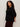 A woman with curly hair stands in profile against a light background, wearing Sanctuary Clothing's ribbed sweater tee in black (extended sizing) paired with burgundy pants.