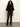 A woman with curly hair wears the Sanctuary Clothing ribbed sweater tee in black, paired with black leather pants, a belt, and tan boots. She stands against a light wall on a gray floor, looking at the camera with a neutral expression.
