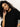 A woman with curly hair, wearing the Sanctuary Clothing ribbed sweater tee in black, poses against a light background, tilting her head and gently touching her face with one hand.