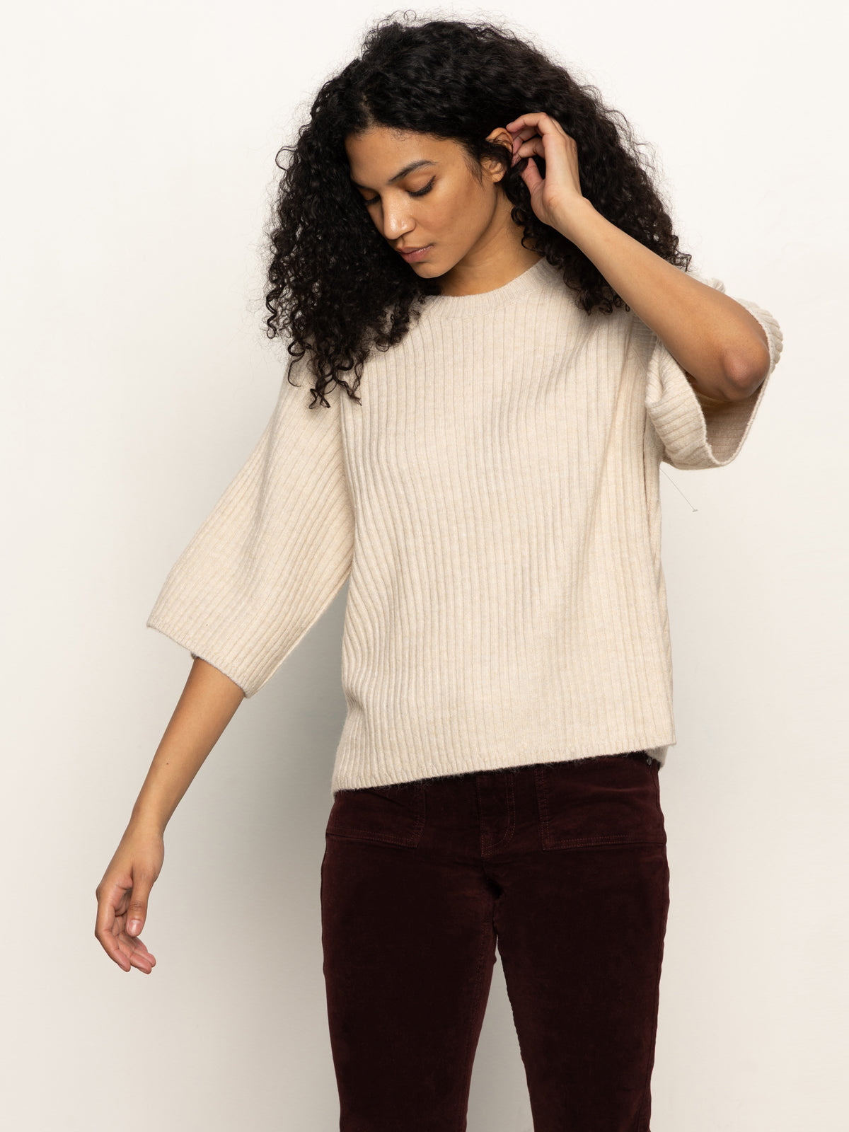 A person with curly hair wears the Sanctuary Clothing ribbed sweater tee in mother of pearl and dark burgundy pants, standing against a plain light background while looking down and touching their hair.