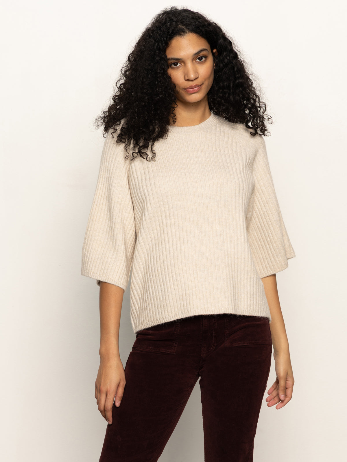 A woman with long curly hair models the Sanctuary Clothing ribbed sweater tee in mother of pearl, featuring wide sleeves, paired with dark burgundy pants against a plain light background.