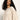 A woman with long, curly dark hair wears the Sanctuary Clothing ribbed sweater tee in mother of pearl, featuring elbow-length sleeves. She stands against a plain, light background and gazes slightly downward with a neutral expression.