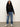 A woman with long curly hair wears the Sanctuary Clothing boheme popover black sweater, blue cuffed jeans, and white sneakers with a black star, standing against a plain light background.