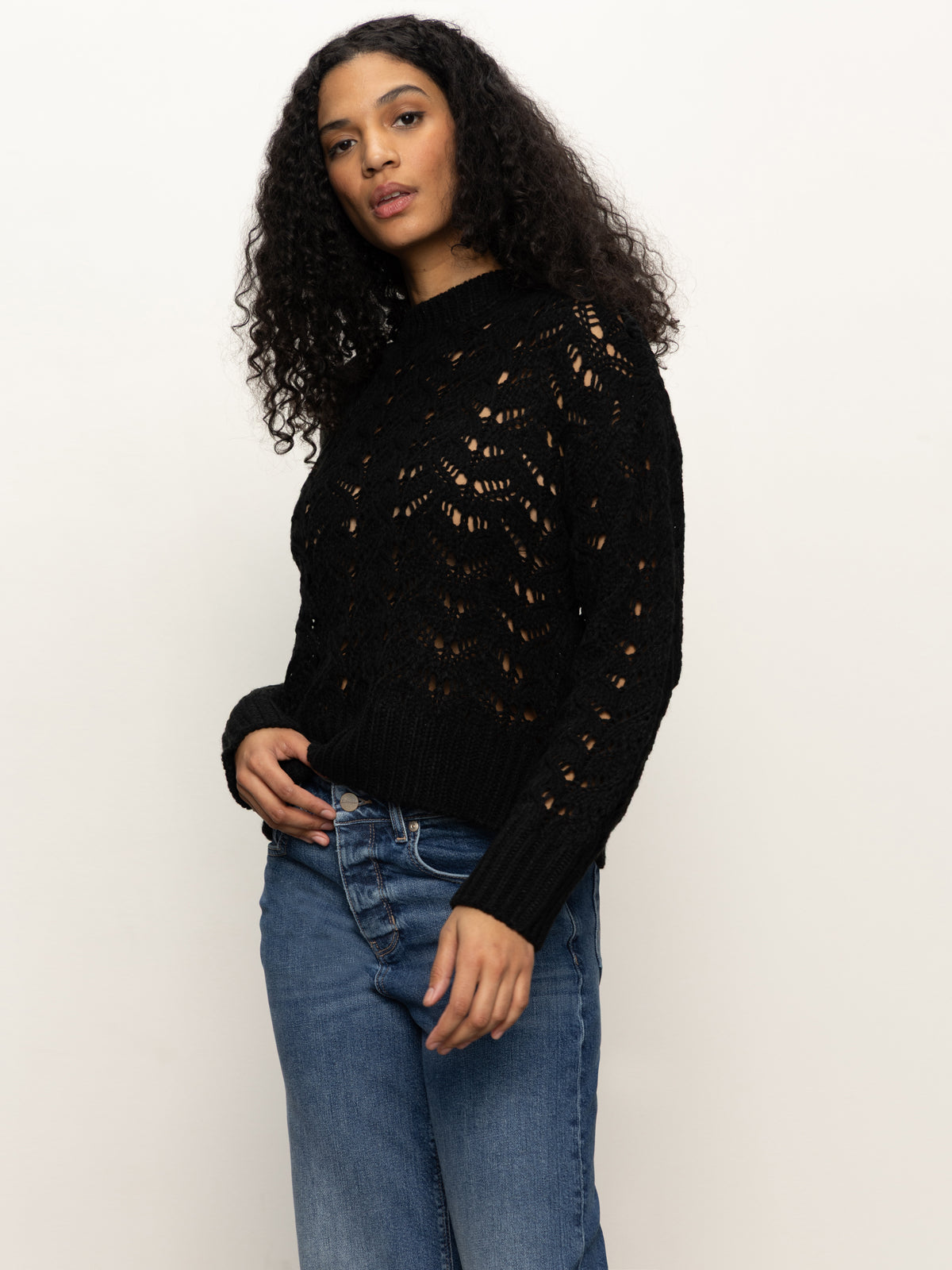 A woman with long curly hair models the Sanctuary Clothing boheme popover black sweater, featuring a lacy knit pattern, paired with blue jeans. She stands against a plain light background, glancing slightly to the side.
