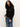 A woman with long curly hair models the Sanctuary Clothing boheme popover black sweater, featuring a lacy knit pattern, paired with blue jeans. She stands against a plain light background, glancing slightly to the side.