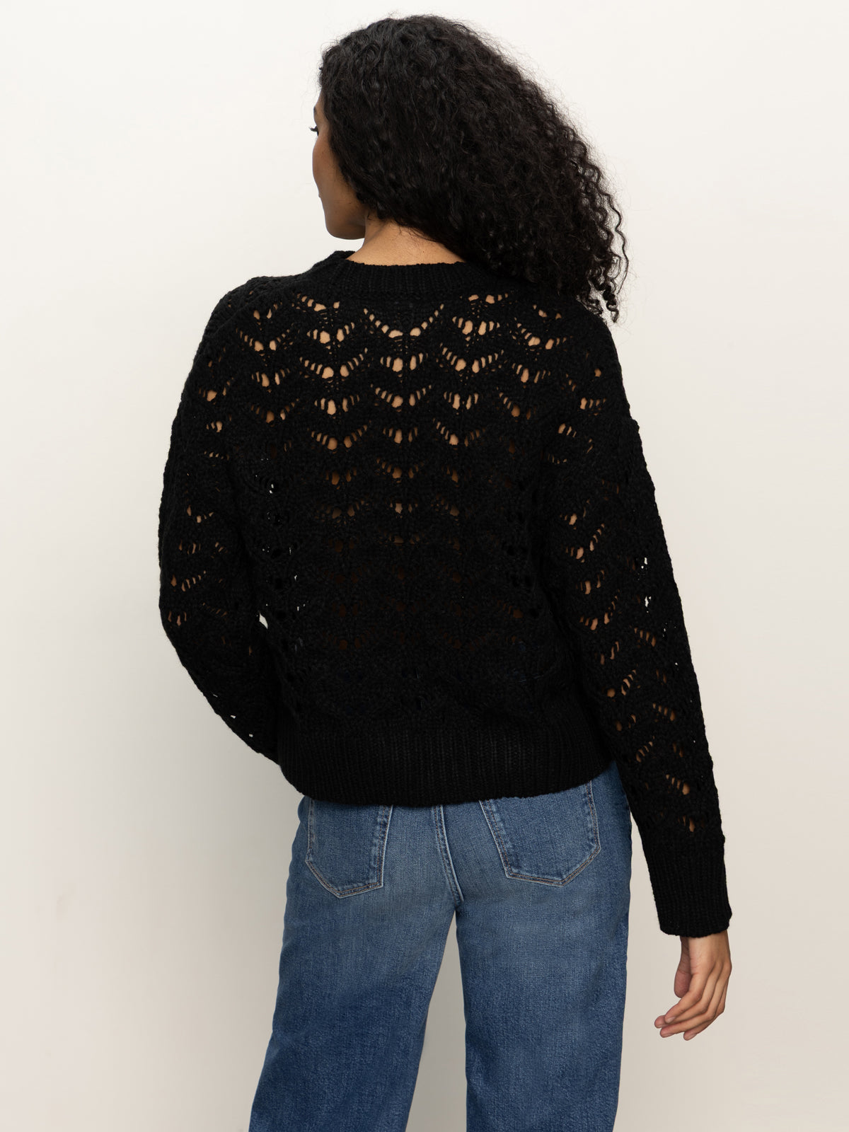 A person with curly hair is seen from behind, wearing Sanctuary Clothing's boheme popover black sweater and blue jeans, standing against a plain light background.