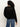 A person with curly hair is seen from behind, wearing Sanctuary Clothing's boheme popover black sweater and blue jeans, standing against a plain light background.