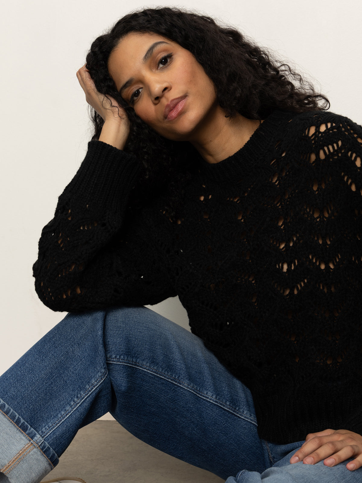 A woman with long curly hair, wearing the Sanctuary Clothing boheme popover black sweater and blue jeans, sits on the floor with one arm on her knee and her head resting on her hand, gazing thoughtfully at the camera.