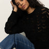 A woman with long curly hair, wearing the Sanctuary Clothing boheme popover black sweater and blue jeans, sits on the floor with one arm on her knee and her head resting on her hand, gazing thoughtfully at the camera.