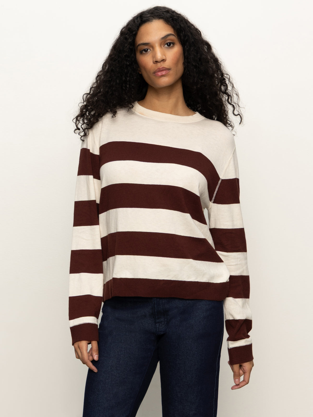 A woman with long curly hair wears the Sanctuary Clothing 24/7 Popover Chicory Light Oat sweater, featuring cream and dark brown horizontal stripes, paired with dark jeans, standing against a plain light background.