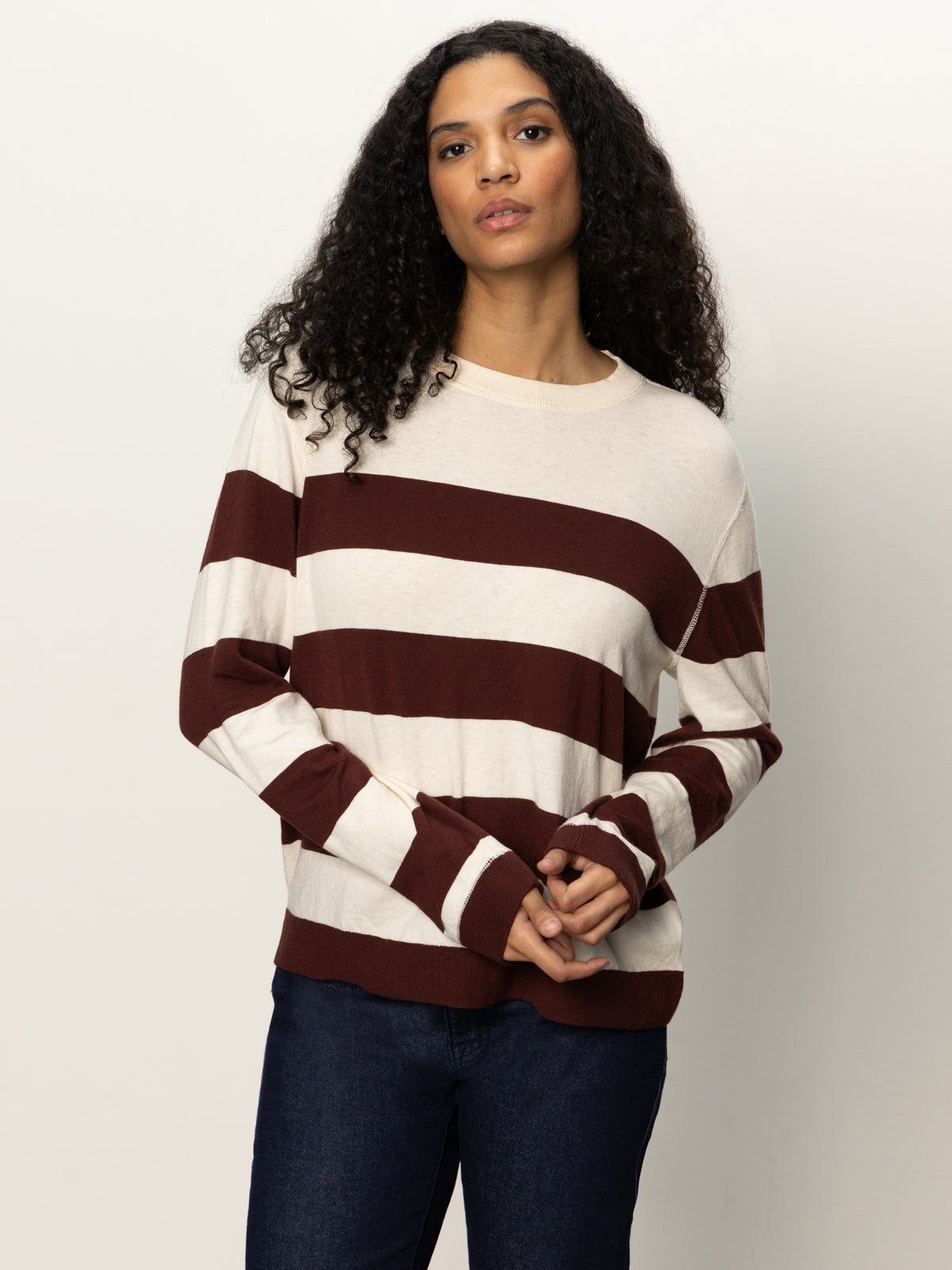 A person with long curly hair wears the Sanctuary Clothing 24 7 popover chicory light oat sweater, featuring wide maroon stripes, paired with dark jeans against a plain light background.