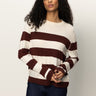 A person with long curly hair wears the Sanctuary Clothing 24 7 popover chicory light oat sweater, featuring wide maroon stripes, paired with dark jeans against a plain light background.