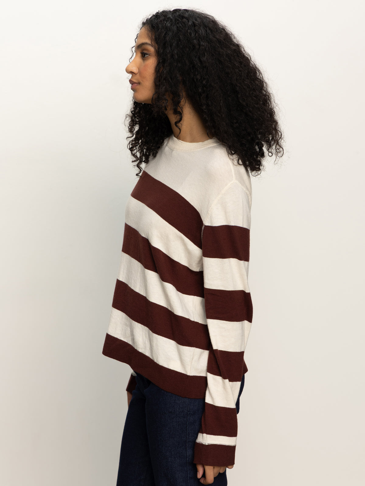 A person with long, curly hair stands sideways against a plain white background, wearing Sanctuary Clothing’s 24 7 popover chicory light oat sweater with dark pants.