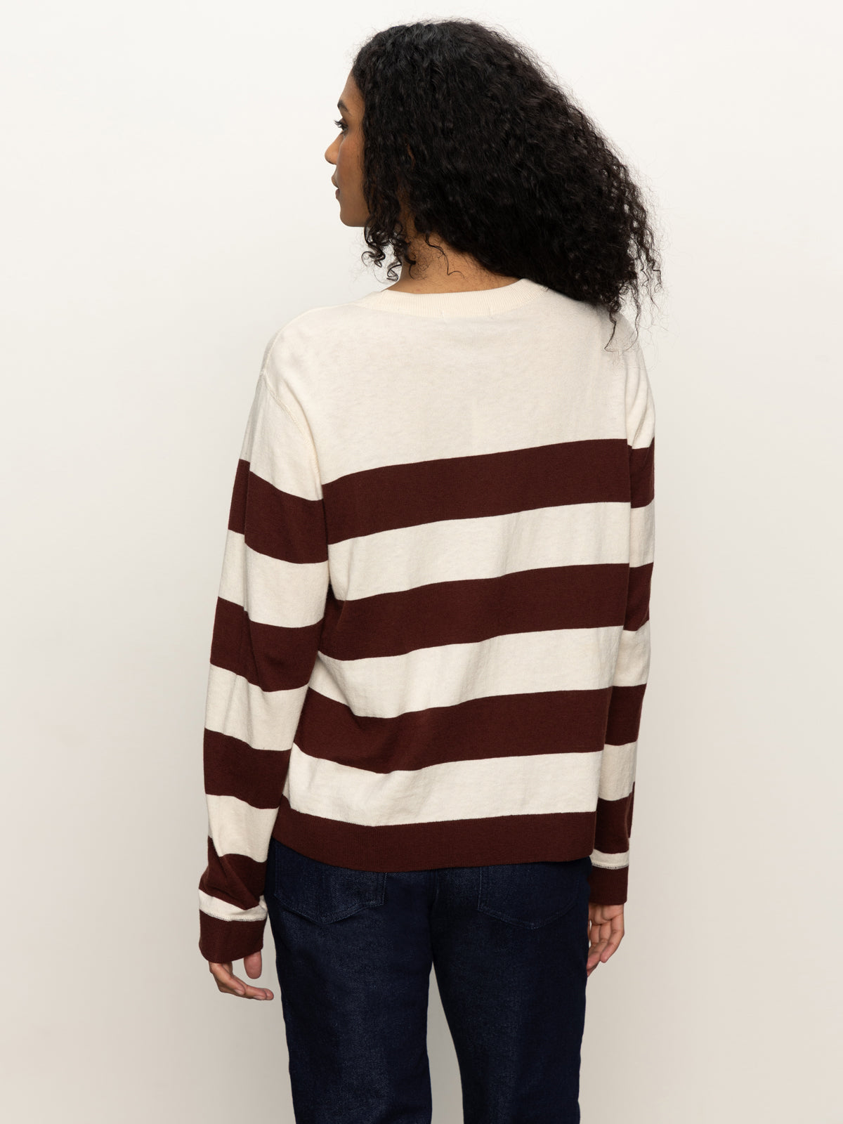 A person faces away, wearing Sanctuary Clothing’s 24 7 Popover in chicory light oat—a cream and brown striped sweater—paired with dark blue jeans, set against a plain light backdrop.