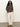 A woman with curly hair stands against a plain background, wearing Sanctuary Clothing's 24 7 Popover French Kitty long-sleeve top, off-white wide-leg pants, and tan ankle boots, with her hands in her pockets.