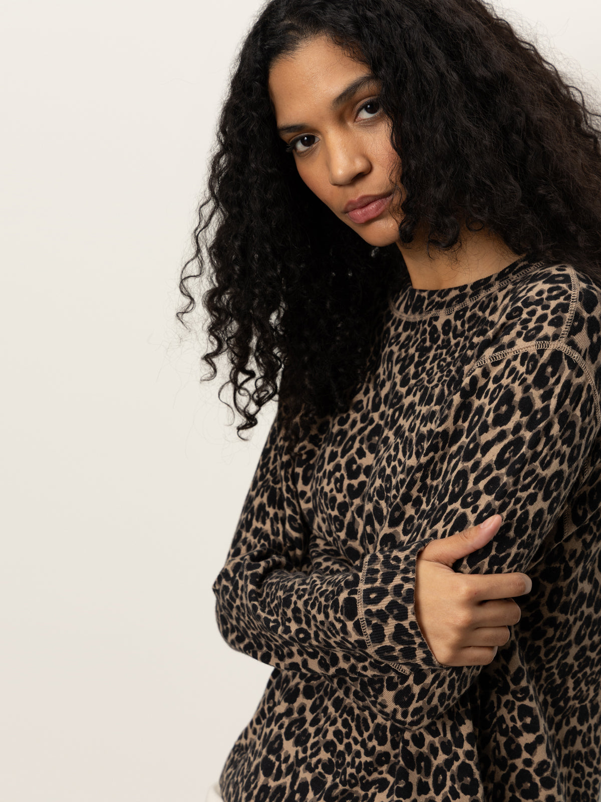 A woman with long, curly dark hair stands against a plain background, wearing the Sanctuary Clothing 24/7 Popover French Kitty top. She looks at the camera with a serious expression, arms crossed gently over her chest.