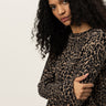 A woman with long, curly dark hair stands against a plain background, wearing the Sanctuary Clothing 24/7 Popover French Kitty top. She looks at the camera with a serious expression, arms crossed gently over her chest.