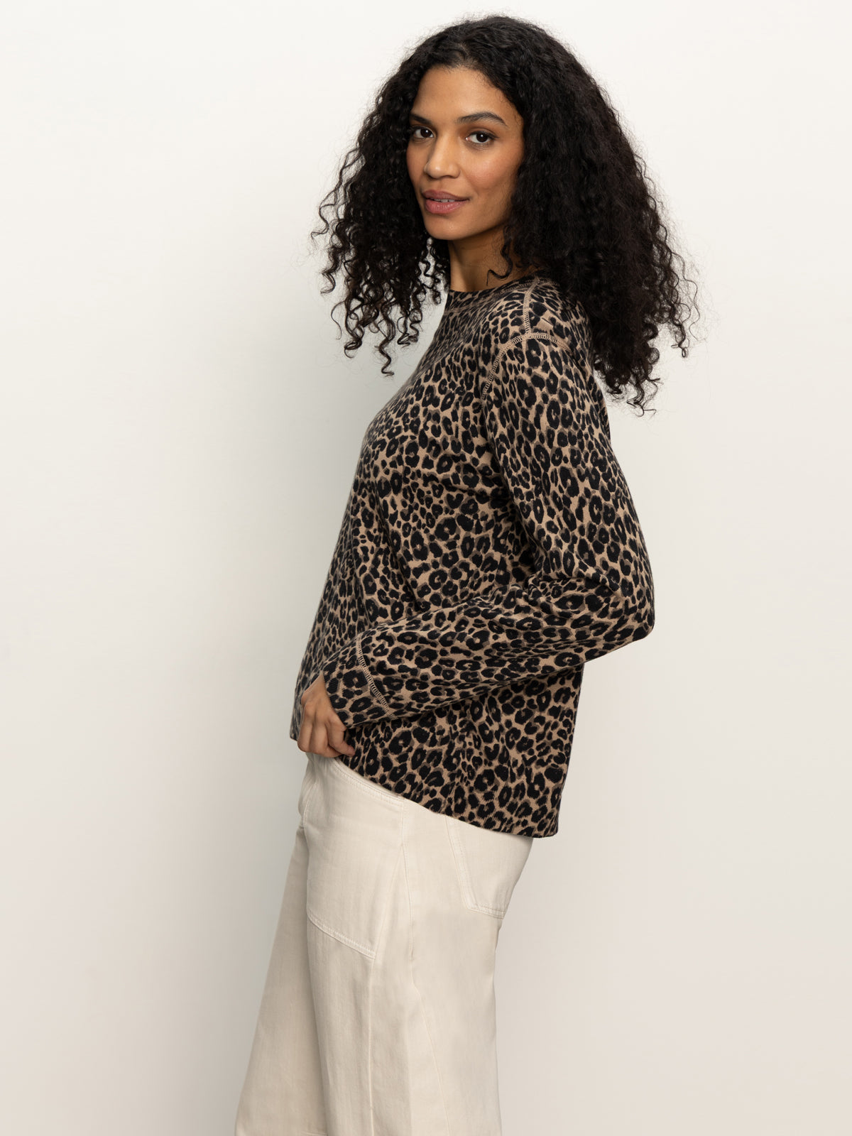 A woman with curly hair stands sideways against a neutral background, wearing the Sanctuary Clothing '24 7 Popover French Kitty' long-sleeve top in brown and black leopard print, paired with light beige pants.