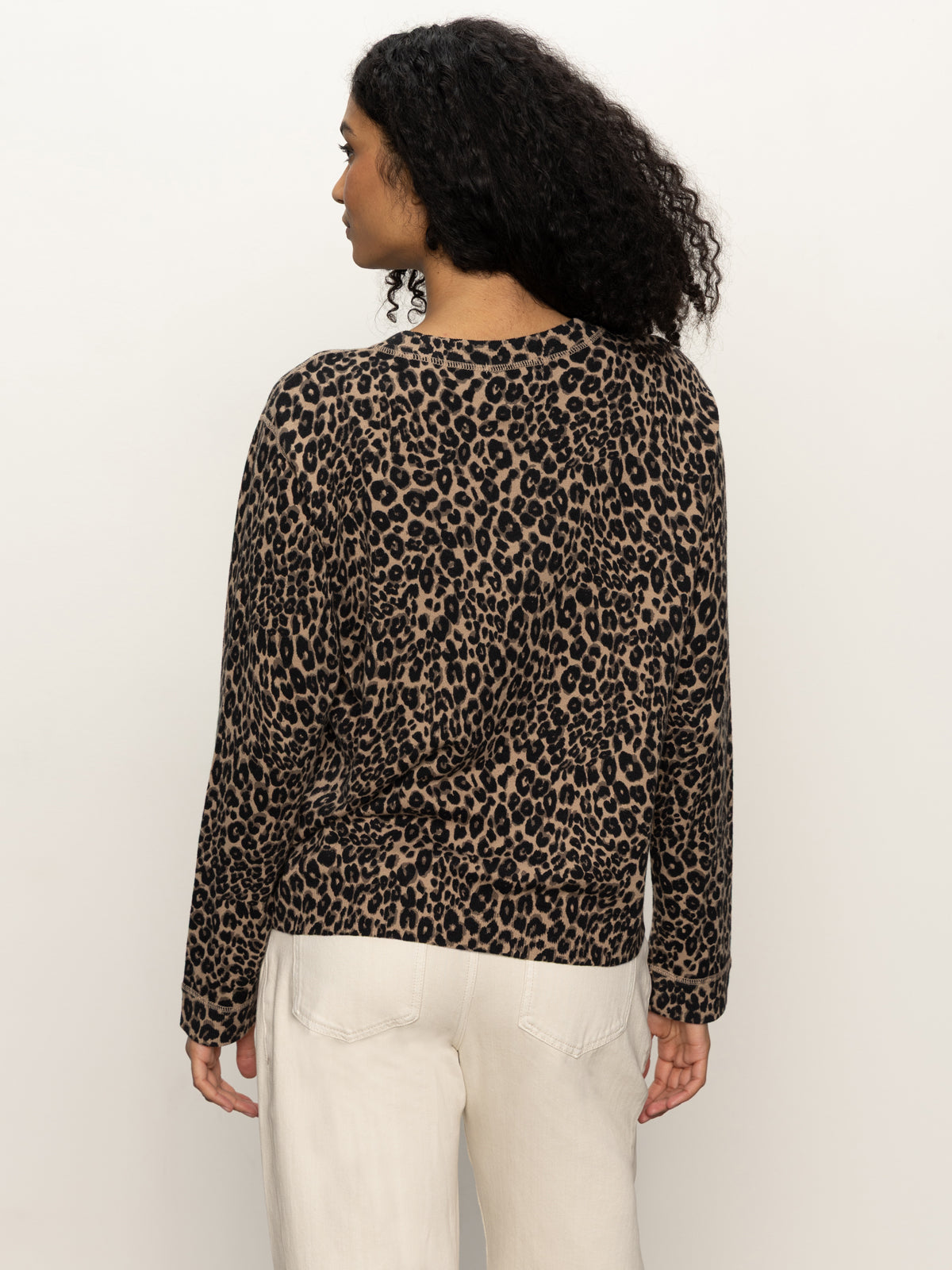 A woman with curly hair is seen from behind wearing the Sanctuary Clothing 24 7 popover french kitty top, featuring a leopard print, with light-colored pants against a plain white background.