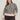 A woman wears the Sanctuary Clothing soft touch popover spots extended sizing sweater in a beige and black leopard print with long sleeves, paired with dark brown pants, standing against a plain light wall and smiling slightly at the camera.
