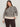 A woman wears the Sanctuary Clothing soft touch popover spots extended sizing sweater in a beige and black leopard print with long sleeves, paired with dark brown pants, standing against a plain light wall and smiling slightly at the camera.