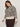 A woman with long brown hair models the Sanctuary Clothing soft touch popover spots extended sizing sweater, featuring a beige and black leopard print with light cuffs and hem, paired with dark brown pants against a light background.
