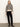 A woman stands against a plain light background, wearing the Sanctuary Clothing "soft touch popover spots" beige sweater with black leopard print, black straight-leg pants, and black ankle boots. She has straight, light brown hair and a neutral expression.