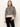 A woman with straight light brown hair and a neutral expression wears Sanctuary Clothing’s soft touch popover spots sweater in beige and black leopard print with long sleeves, paired with black pants, standing against a plain light background.