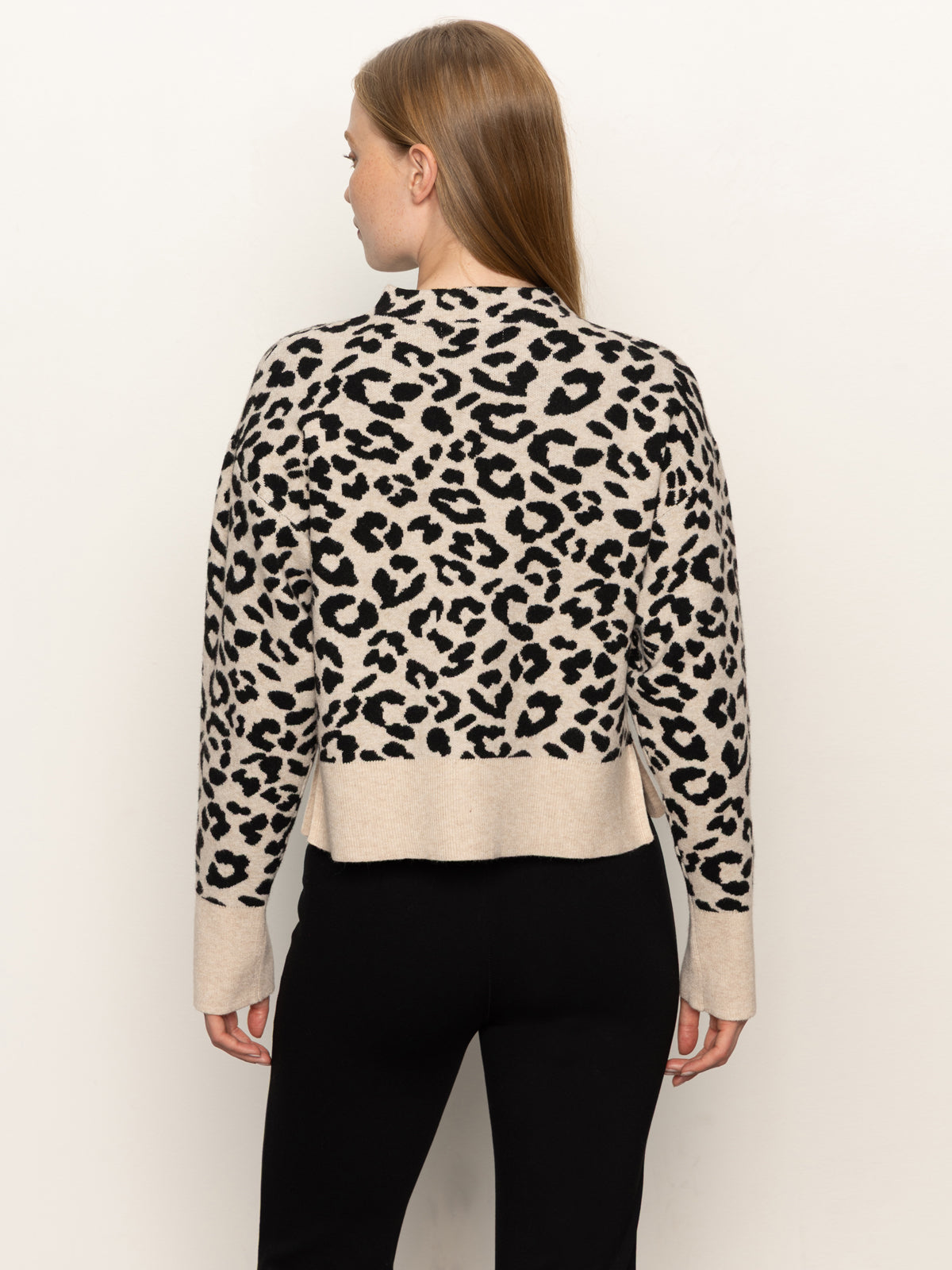 A woman with straight, light brown hair is seen from the back modeling Sanctuary Clothing’s soft touch popover spots sweater—beige with a black leopard print—paired with black pants, standing against a plain light background.