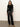 A woman with loose, wavy hair and a neutral expression wears the Sanctuary Clothing long sleeve shoulder twist top in black, paired with high-waisted black leather pants and tan pointed-toe boots against a light background.
