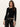 A young woman with long brown hair wears the Sanctuary Clothing long sleeve shoulder twist top in black, paired with black leather pants and a studded belt, standing against a plain light-colored wall.