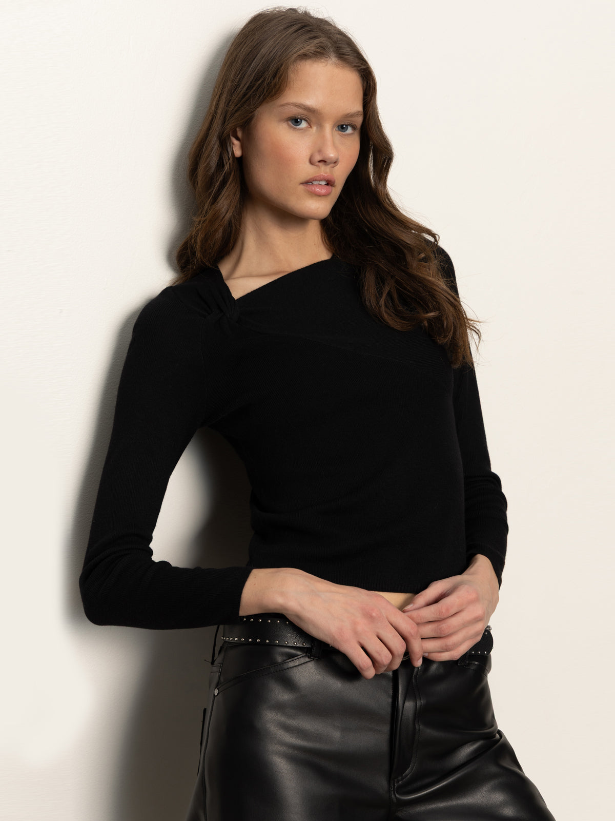 A woman with long brown hair wears the Sanctuary Clothing long sleeve shoulder twist top in black, paired with high-waisted black leather pants featuring stud details, as she poses against a light-colored wall.