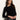 A woman with long brown hair wears the Sanctuary Clothing long sleeve shoulder twist top in black, paired with high-waisted black leather pants featuring stud details, as she poses against a light-colored wall.