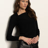 A woman with long brown hair wears the Sanctuary Clothing long sleeve shoulder twist top in black, paired with high-waisted black leather pants featuring stud details, as she poses against a light-colored wall.