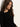 A young woman with long wavy brown hair, wearing the Sanctuary Clothing long sleeve shoulder twist top in black, stands against a light-colored wall and looks down calmly.