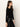 A woman with long brown hair wears the Sanctuary Clothing long sleeve shoulder twist top in black, styled with black leather pants and a studded belt, standing in profile against an off-white background.
