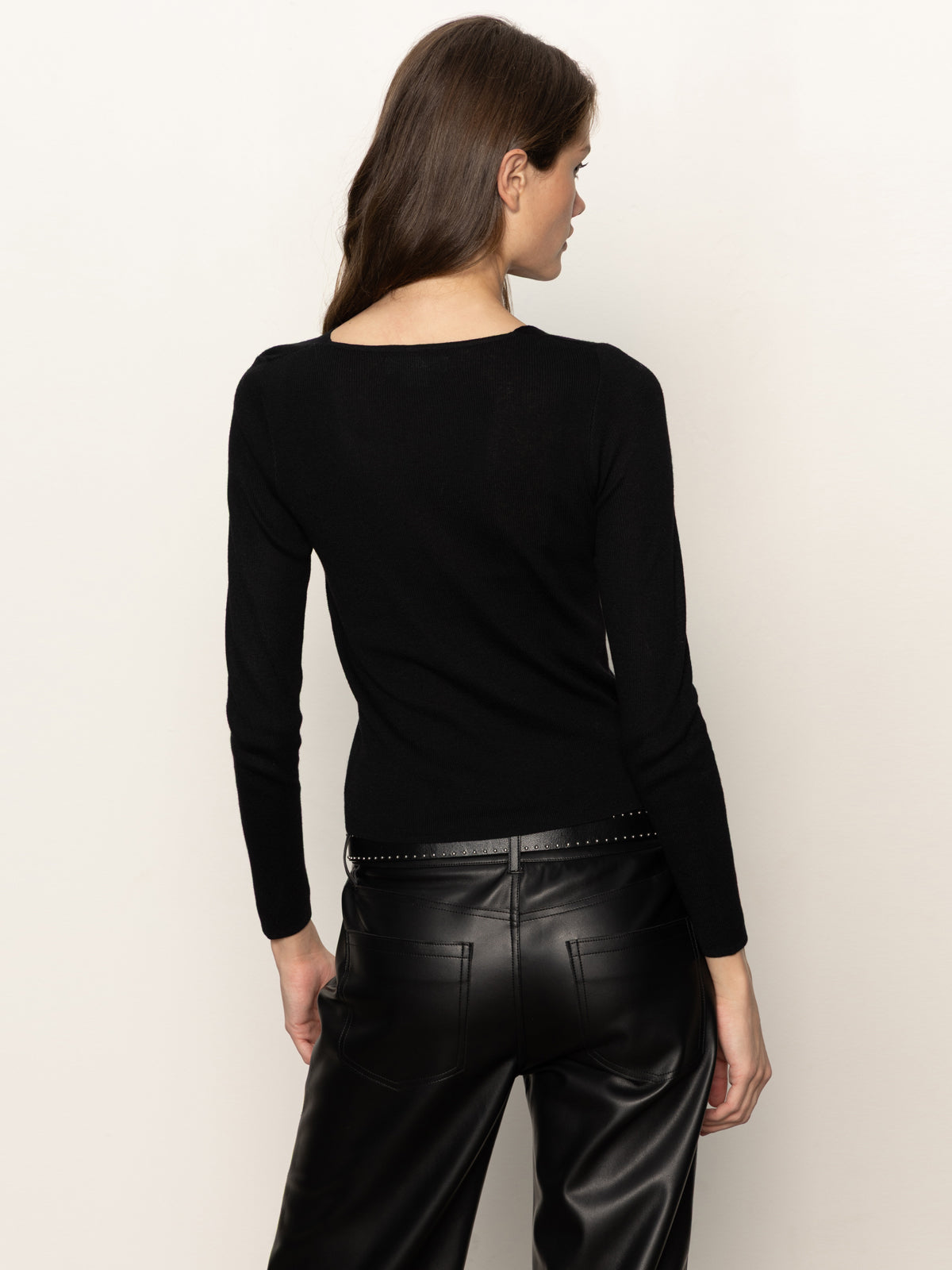 A woman with long brown hair faces away, wearing the Sanctuary Clothing long sleeve shoulder twist top in black and high-waisted black leather pants with silver studs, set against a plain light background.