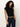 A woman with curly hair wears Sanctuary Clothing's asymm shoulder twist top in black, paired with dark high-waisted jeans and a leopard print belt, standing against an off-white background with one hand in her pocket.