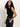 A woman with long curly hair wears the Sanctuary Clothing asymm shoulder twist top in black, styled with dark blue jeans and a leopard print belt, standing against a plain light background.