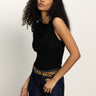 A woman with long curly hair wears the Sanctuary Clothing asymm shoulder twist top in black, styled with dark blue jeans and a leopard print belt, standing against a plain light background.