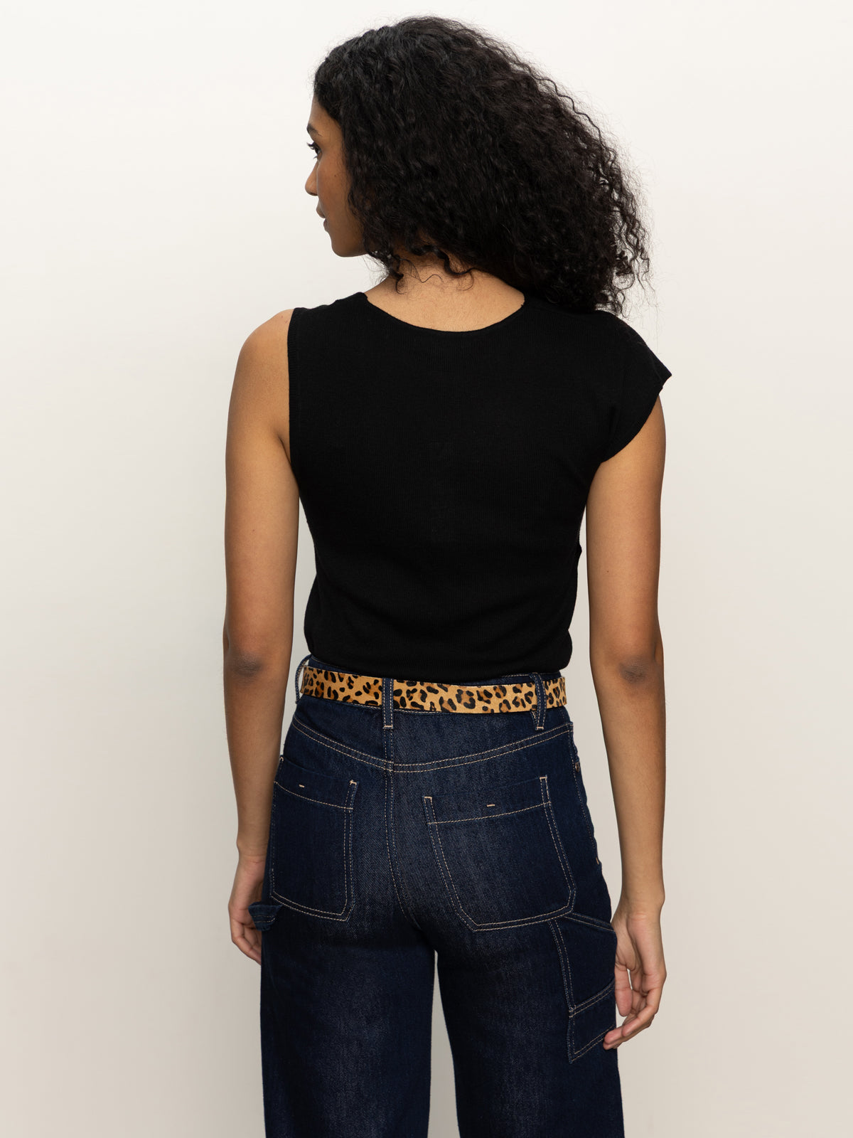 A person with curly hair wears the Sanctuary Clothing asymm shoulder twist top in black, dark blue jeans, and a leopard print belt, standing with their back to the camera against a plain light background.