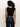 A person with curly hair wears the Sanctuary Clothing asymm shoulder twist top in black, dark blue jeans, and a leopard print belt, standing with their back to the camera against a plain light background.