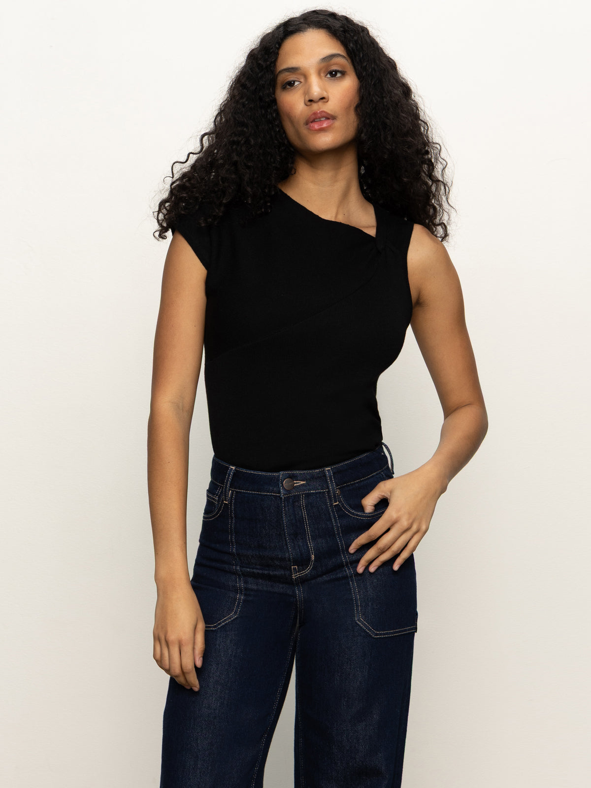 A woman with long curly hair wears the Sanctuary Clothing asymm shoulder twist top in black and high-waisted dark blue jeans, standing against a plain light background with one hand on her hip.