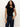 A woman with long curly hair wears the Sanctuary Clothing asymm shoulder twist top in black and high-waisted dark blue jeans, standing against a plain light background with one hand on her hip.