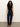 A woman with long curly hair wears a black sleeveless top, high-waisted dark blue jeans with white stitching, and tan heeled boots, standing against a plain light-colored background.