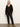 A woman stands against a light wall wearing the meow cardi kitty camo by Sanctuary Clothing, featuring a brown and black leopard print with large buttons, paired with black leather pants and brown suede ankle boots. Her hair is straight and she looks neutral.