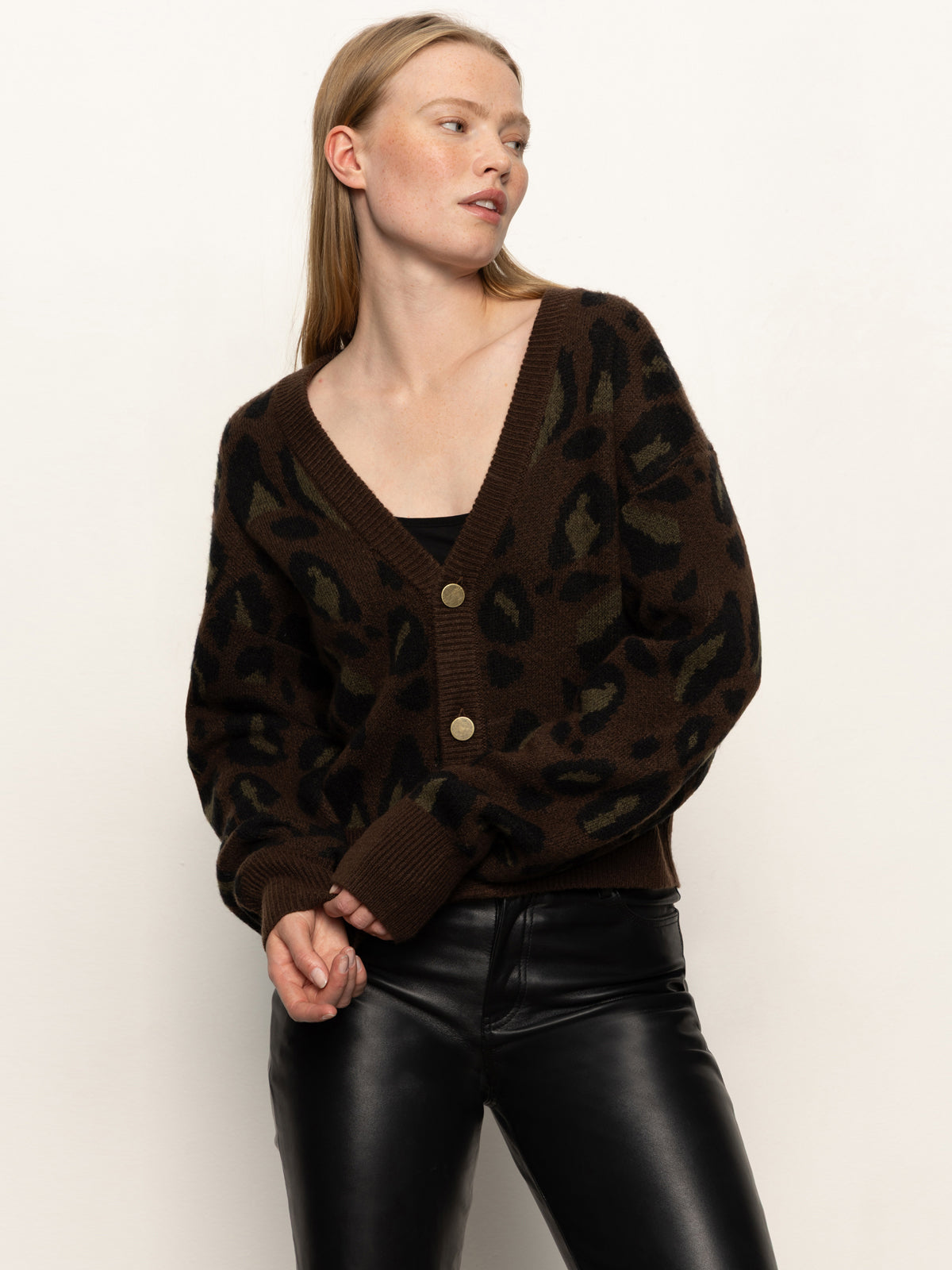 A woman models the Sanctuary Clothing meow cardi kitty camo, a dark brown leopard print cardigan with large buttons, over a black top and black leather pants, standing against a plain background and looking to her left.