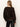 A person with long light brown hair is shown from the back, wearing Sanctuary Clothing’s meow cardi kitty camo sweater and black leather pants, standing against a plain light-colored wall.