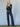 A woman stands with arms crossed, wearing the Sanctuary Clothing ekara slim wide jean in indigo dark rinse wash, paired with a black long-sleeve top, black belt, and heeled boots against a plain background.