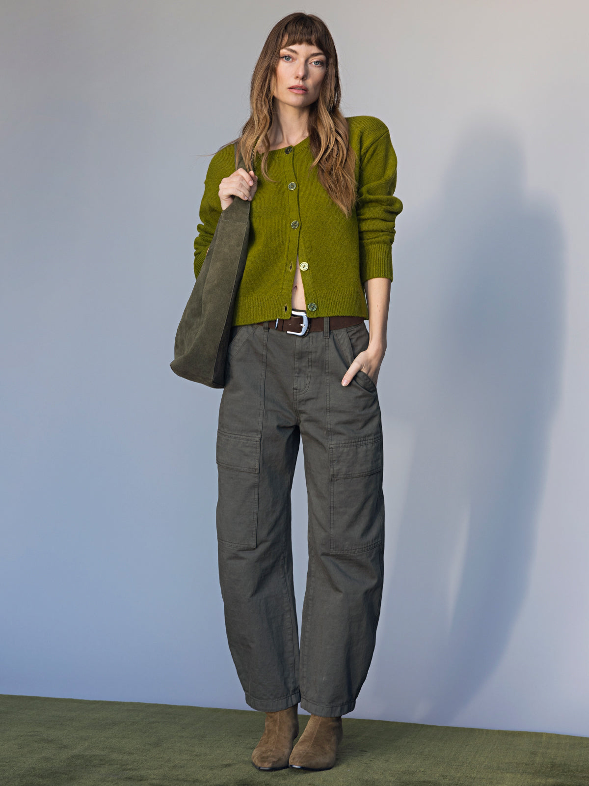 A woman with long brown hair and bangs poses confidently against a neutral background, carrying the Sanctuary Clothing endless large hobo bag in dark olive. She wears a matching cardigan, gray cargo pants, and brown ankle boots.